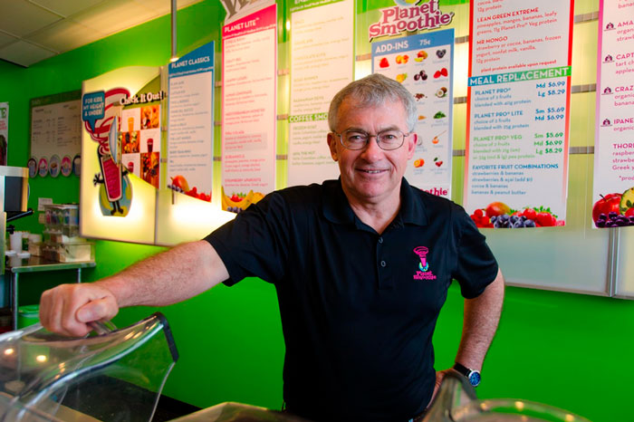 Blair Ritchey, the North Texas Area Developer of Planet Smoothie Franchises, stands smiling in a planet smoothie uniform in front of a bright green, well lit wall