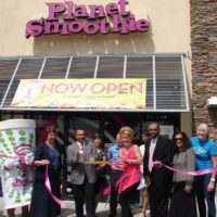 At a planet smoothie grand opening a group of business men and women cut a pink ribbon