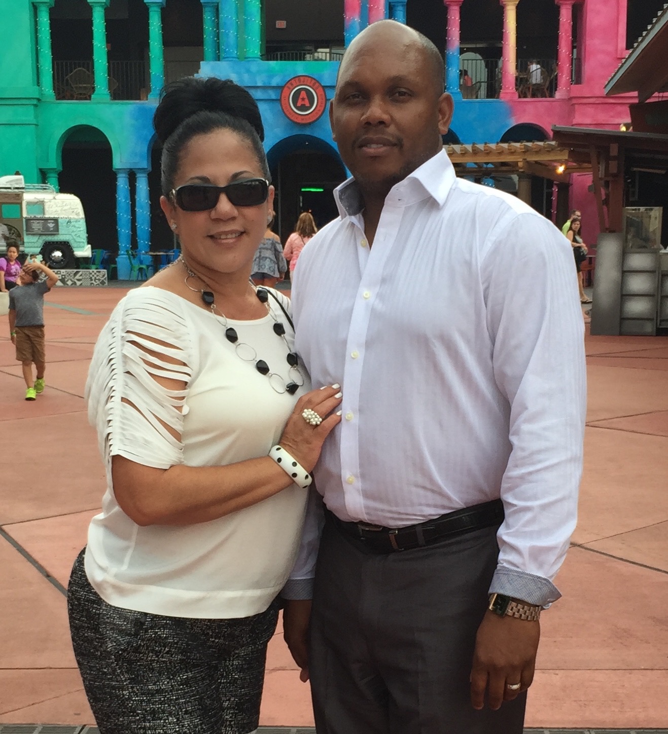 Tyrone Forbes, Planet Smoothie Franchise owner, stands with his wife in front of a bright green, blue, and pink background in a button up shirt and a smile on his face while his wife rests her hand on his chest and smiles at the camera